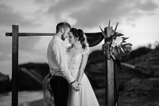 young couple groom and bride with a bouquet in the evening on the beach near the wedding arch