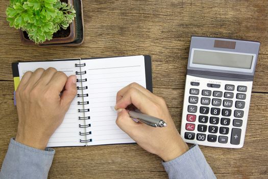 Hands using pen write a notepad with calculate Calculator, and green plant on wooden background.