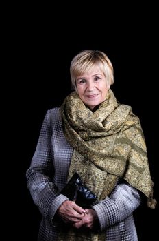 Portrait of an elderly woman in a draped coat with a thrown shawl on her shoulders and a clutch bag. Black background.