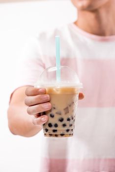 Hand holding light brown creamy bubble tea and black tapioca pearls in plastic cups on table.