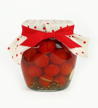 Close up of one glass jar of pickled small red cherry tomatoes with linen canvas lid decoration and red ribbon over white background, low angle side view