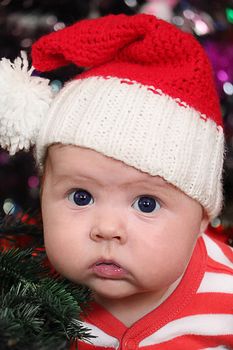 Portrait of astonished newborn baby with beautiful blue eyes  in santa hat and striped jacket on blurred background and christmas tree needles. 