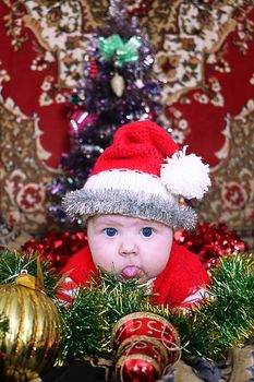 Newborn naughty Santa baby with amazing blue eyes dressed  in red santa hat and jacket shows tongue out laying on tinsel and christmas decorated background, Christmas tree behind of him. New year card