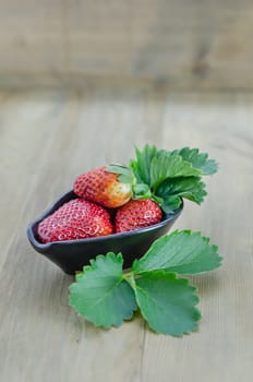 fresh strawberry fruits in black bowl with green leaves over wooden background