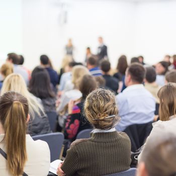 Business and entrepreneurship symposium. Female speaker giving a talk at business meeting. Audience in conference hall. Rear view of unrecognized participants making notes in audience.