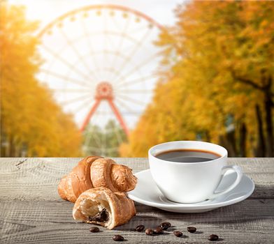A cup of coffee with croissants on a wooden table. Grains of coffee on the table. Blurred background of a ferris wheel in autumn in the park.