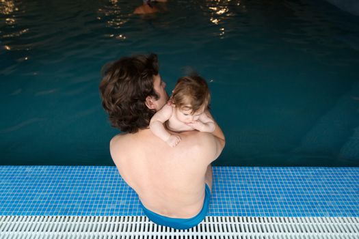 Family of father and his little daughter sitting at edge of swimming pool. Young father and his little cute newborn baby together in spa hotel. Dad holding child adorable girl on arms. Family relaxing.