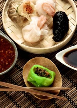 Assorted Dim Sum in Bamboo Steamed Bowl and Vegetarian Yasai on Wooden Plate with Red Chili and Soy Sauces and Chopsticks closeup on Straw Mat background