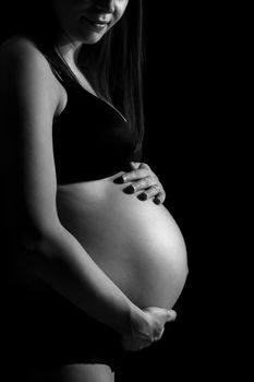 Pregnant woman caressing her belly isolated on black monochrome background with copy space for text. Unrecognizable pregnant woman with hands over tummy. Black and white photo.