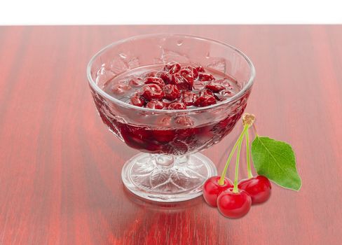 Cherry jam with whole fruits in the glass dessert stem bowl and three fresh cherries beside  on a red wooden table
