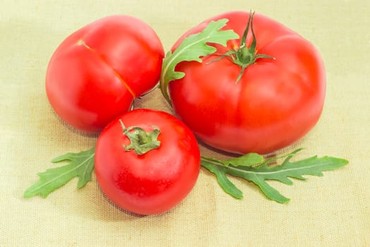 Three ripe red tomatoes different sizes and arugula leaves closeup on a sackcloth
