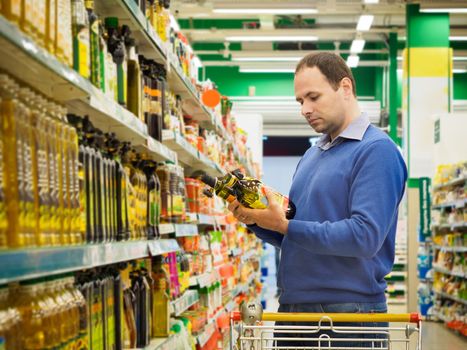 Man making choise about two bottles of oil from shelf in supermarket. Consumerism concept