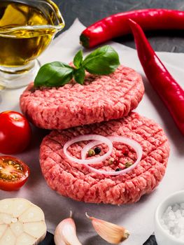 Close up view of two raw meat steak cutlets for burger with vegetables, spices, fresh basil, olive oil. Making homemade burger. Vertical.