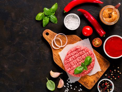 Raw beef meat steak cutlet for burger on wooden cutting board with spices, vegetables, sauces ketchup and mustard on black concrete background. Making homemade burger. Top view or flat lay. Copy space