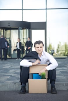 Fired business man sitting frustrated and upset on the street near office building with box of his belongings. He lost work