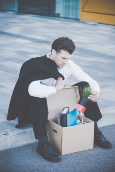 Fired business man sitting frustrated and upset on the street near office building with box of his belongings. He lost work