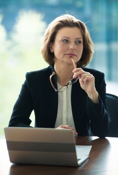 Mature business woman with glasses sitting at meeting table in office with laptop