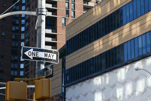 ONE WAY road sign points left on a New York City street, with high rise buildings beyond