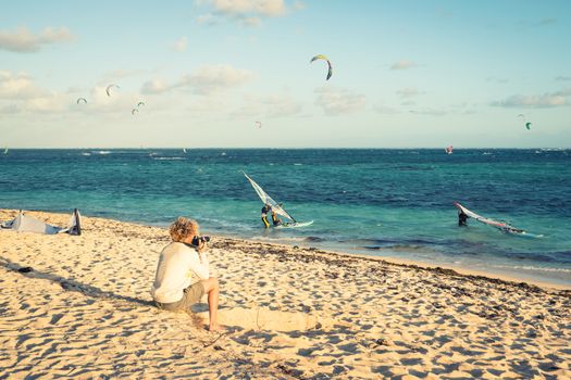 Travel photographer  taking pictures sitting on the beach during event of kitesurfing