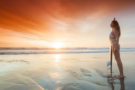 A beautiful young slim sporty woman in bikini with a surfboard is standing at ocean beach at sunset