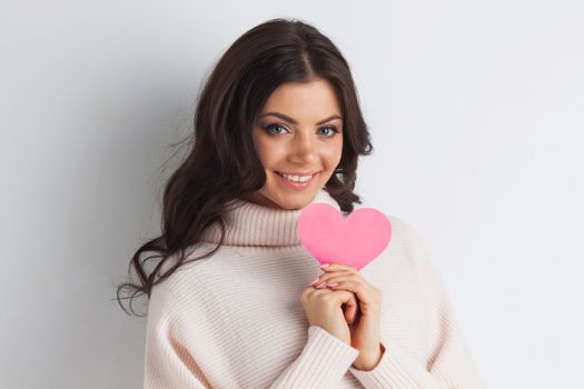 Beautiful young brunette woman holding pink paper heart, looking at camera