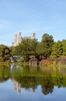 Turtle Pond, Central Park, on a sunny autumn day. Surrounding trees and plants reflect in the still water. The rooftop of the Beresford Hotel can be seen outside the grounds.