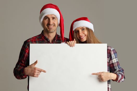 Happy couple in Santa hats holding blank banner and pointing at copy space