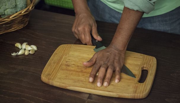 An afro-american man s hand is pressing with a knife garlic on a wooden board.