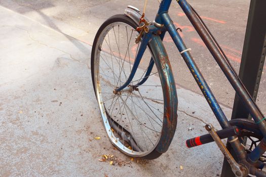 Broken front wheel with loose tyre on an abandoned rusty bicycle, locked to a metal pole.
