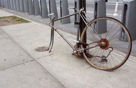 Broken, rusty bicycle with missing front wheel and dropped chain locked to a metal pole on a sidewalk
