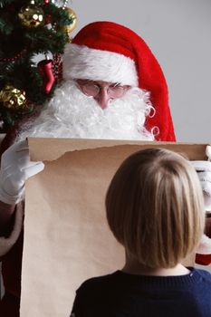 Santa Claus with with list or nice or naugthy list paper with small boy near christmas tree