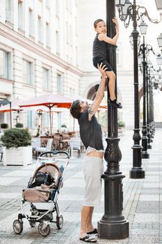 A handsome father is holding up his young boy on a street pillar.