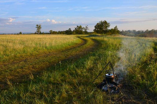 a kettle hangs over the fire, in which water for coffee is heated. A summer tour in nature. Landscape with grass field and sky