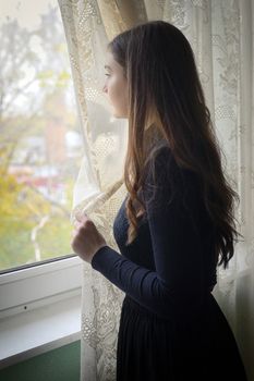 Young girl in black vintage dress standing near window 