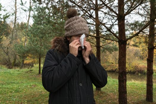 Young man wearing jacket suffering from cold and holding handkerchief on nose. Male in knitted hat standing in the wood blowing his nose. Autumn illness and flu season concept