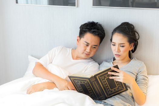 Young Asian Couple laid in a bed reading a book in bedroom of contemporary house for modern lifestyle concept