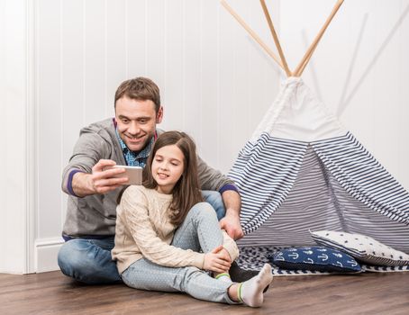 Father and daughter taking selfie playing at home with constructed wigwam