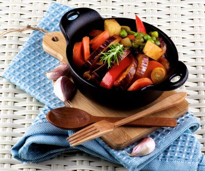 Delicious Homemade Colorful Vegetables Ragout with Eggplant, Carrots, Potatoes, Leek, Bell Pepper and Green Pea in Black Iron Cast with Wooden Spoon and Fork closeup on Blue Napkin