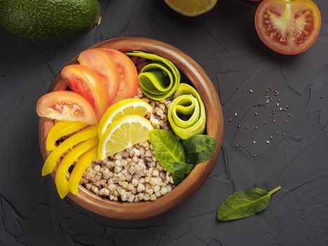 Top view of vegetarian buddha bowl with green buckwheat, spinach, avocado, tomatoes and yellow sweet pepper paprika on dark concrete background with copy space