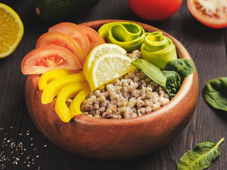 Close up view of vegetarian buddha bowl with green buckwheat, spinach, avocado, tomatoes and yellow sweet pepper paprika on dark wooden table with copy space