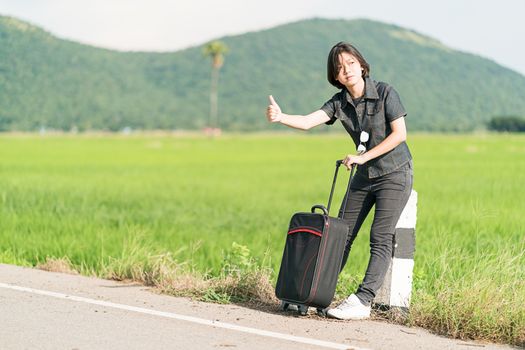 Young asian woman short hair and wearing sunglasses with luggage hitchhiking along a road and thumbs up in country road Thailand