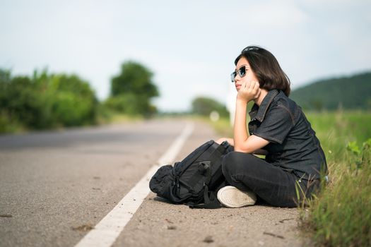 Young asian woman short hair and wearing sunglasses sit with backpack hitchhiking along a road wait for help in country road Thailand