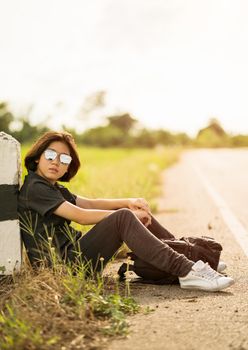 Young asian woman short hair and wearing sunglasses sit with backpack hitchhiking along a road wait for help in country road Thailand