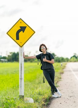 Young asian woman short hair and wearing sunglasses with backpack hitchhiking along a road in countryside Thailand