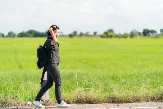 Young asian woman short hair and wearing sunglasses with backpack hitchhiking along a road in countryside Thailand