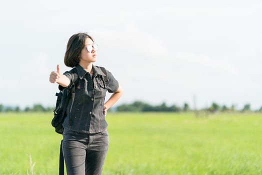 Young asian woman short hair and wearing sunglasses with backpack hitchhiking along a road in countryside Thailand