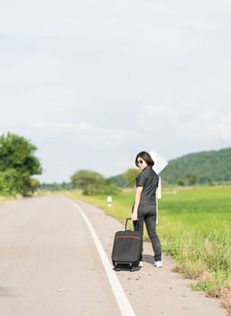 Young asian woman short hair and wearing sunglasses with luggage hitchhiking along a road in countryside Thailand