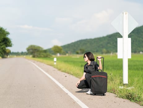 Young asian woman short hair and wearing sunglasses with luggage hitchhiking along a road in countryside Thailand