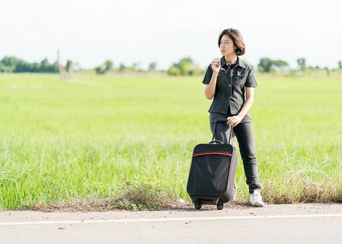 Young asian woman short hair and wearing sunglasses with luggage hitchhiking along a road in countryside Thailand