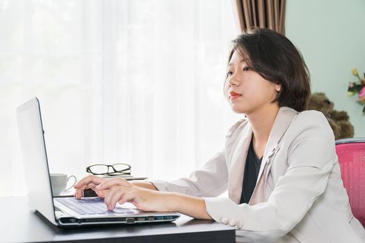 Young asian woman short hair in smart casual wear working on laptop while sitting near window in home office